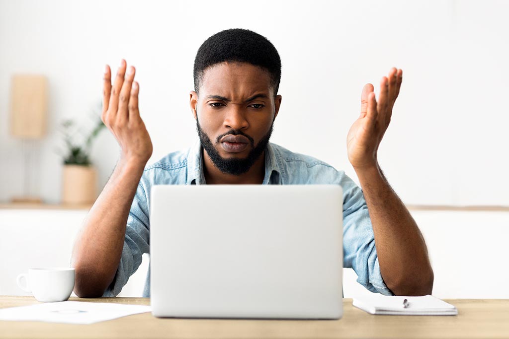 A frustrated man raises his hands in exasperation while working on a laptop at his desk.